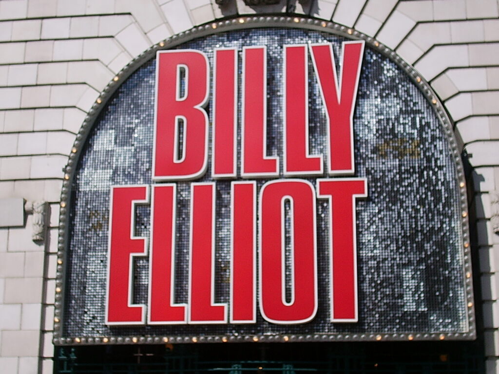 Alt text: A photo of the “Billy Elliot the Musical” theater sign taken in 2007. The sign features bold red lettering on a white background mounted above the entrance, promoting the stage production of Billy Elliot the Musical.