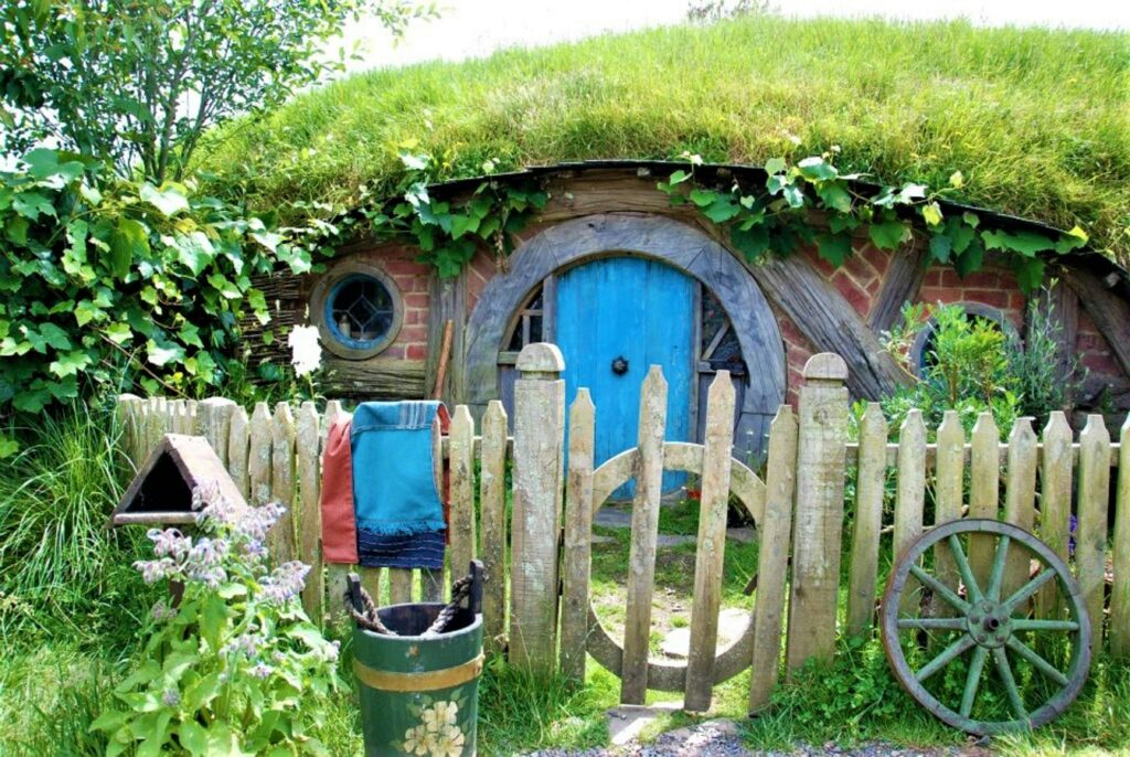 A hobbit-style cottage built into a grassy hillside with a round turquoise-blue door and matching circular windows, framed by weathered red brick and curved wooden beams. A grass-covered roof drapes with ivy above the doorway, and a rustic wooden picket fence with a round gate sits in the foreground, decorated with a woven blanket, a daisy-painted bucket, a small birdhouse, purple wildflowers, and an old cartwheel leaning against the fence.