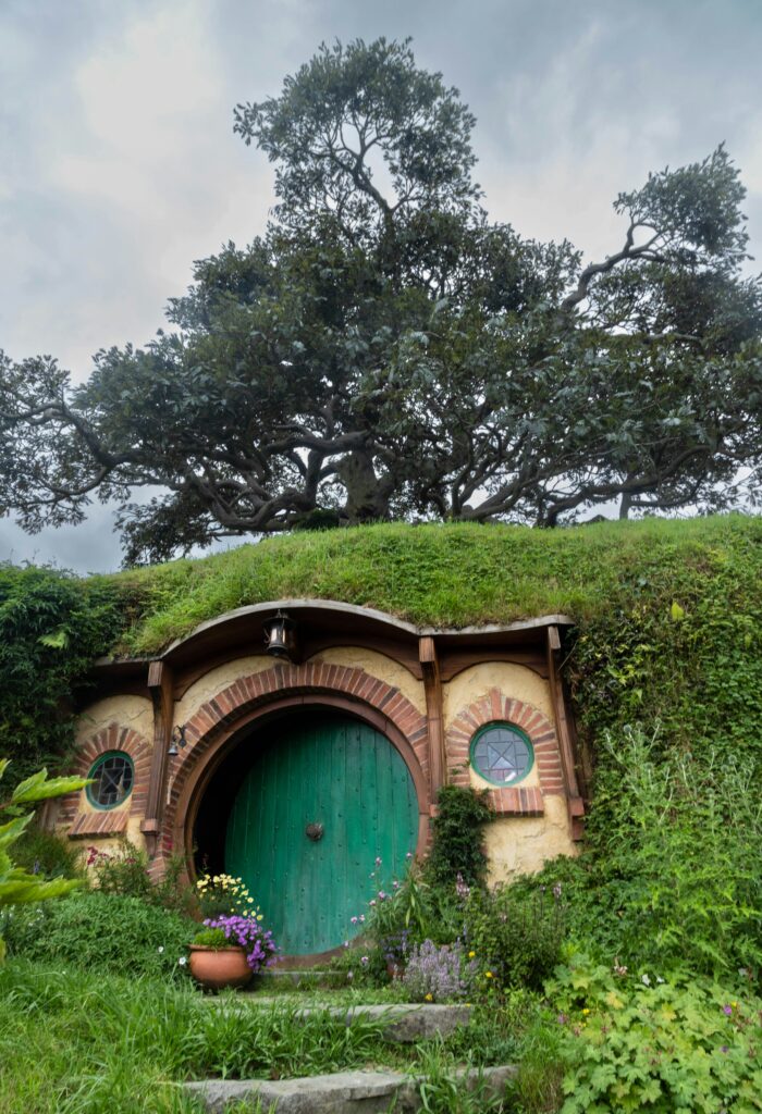 A round, arched wooden door painted green, set into a grassy hillside and framed by lush greenery, ferns, and overhanging plants, with a stone pathway leading up to it, evoking a cozy hobbit hole.