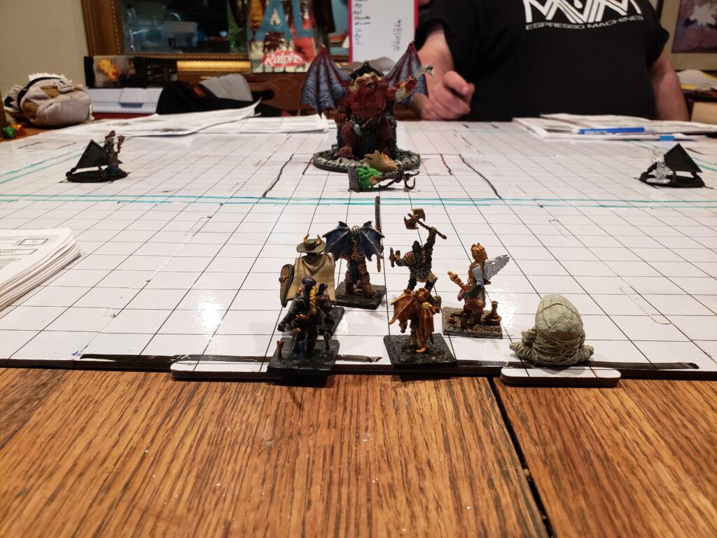 abletop miniatures arranged on a grassy terrain board with trees, rocks, and rulers used for measuring movement. Multiple players sit around the table, discussing tactics and moving units together, illustrating cooperative tabletop play where shared strategy matters more than resource tracking.