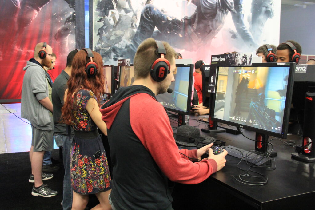 Crowds at RTX 2015 gather around a gaming booth where players at multiple monitors test fast‑paced shooters. Bright screens show continuous gunfire and rapid action, illustrating the nonstop combat pacing often associated with unlimited ammo gameplay in cooperative and competitive settings.