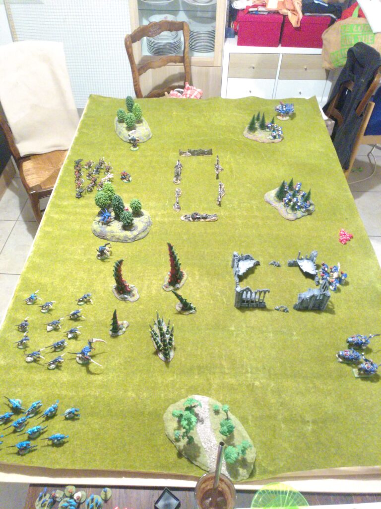 A Warhammer 40,000 tabletop battle between Tyranids and Space Marines is set up on a large terrain board with buildings, rubble, and miniatures positioned for combat. Two players stand on opposite sides of the table, measuring distances and moving units. The scene shows brightly painted Tyranid creatures advancing toward Space Marine squads, with dice and templates visible around the battlefield.
The caption reads:
Warhammer 40K doesn’t track ammunition for most weapons, allowing units to fire freely every turn. This creates a functional version of the Unlimited Ammo Rule through abstraction, supporting fast pacing and cinematic combat.
