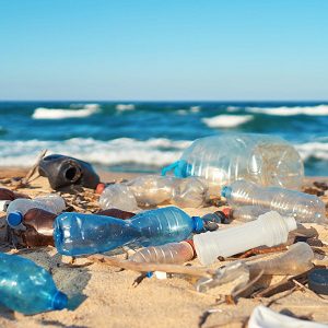 A sandy beach scattered with plastic waste including bottles, containers, and assorted debris, with ocean waves rolling in under a clear blue sky, highlighting the impact of marine pollution.